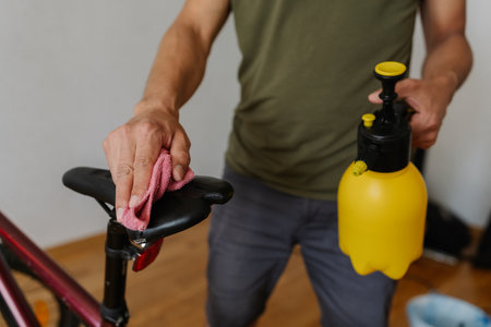 Vertical cropped shot of cyclist cleaning saddle of mountain bike using spray bottle and cloth, ensuring spotless and ready for next ride at home, close-up. Concept of bicycle maintenance and service.の写真素材