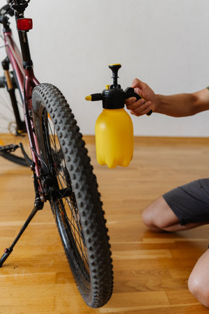 Close-up of cyclist using yellow handheld water sprayer to clean dusty dirty tire wheel of mountain bike at home. Concept of bike maintenance, service and cleaning practicesの写真素材
