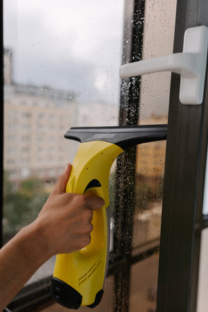 Close-up of male hand using cordless vacuum cleaner for cleaning window, wiping away water drops on rainy day, ensuring spotless and hygienic indoor environment. Concept of household cleanliness.の写真素材