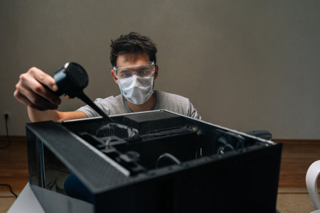 Front view of technician male wearing face mask and safety glasses cleaning inside of computer case...