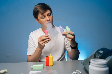 Portrait of man sitting at desk with scattered cash, smoking electronic heated tobacco device,...