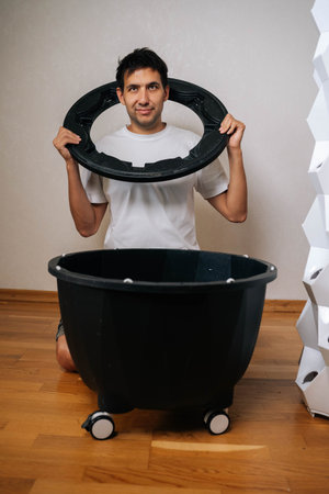 Experienced gardener assembling components of hydroponic system, holding circular part over black container on wheels, with vertical hydroponic garden in background, looking at camera.の写真素材