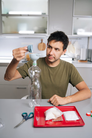 Vertical portrait of young man pouring organic fertilizer through funnel into plastic bottle,...
