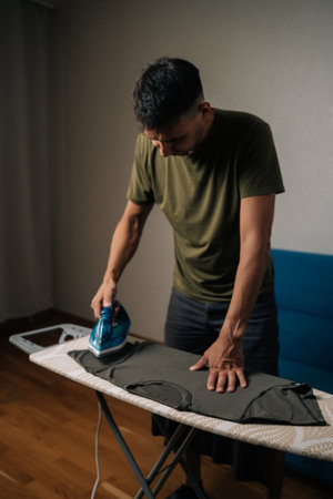 Vertical shot of focused man ironing clothes on ironing board in cozy living room, managing...