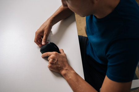 Top view of technician male cleaning disassembled vertical mouse with disinfectant wipe sitting at white table, ensuring hygiene and optimal performance in professional setting, close-up.の写真素材