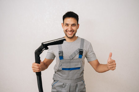 Portrait of cheerful professional cleaning service employee in overall holding washing vacuum cleaner, getting ready to cleaning in living room, smiling looking at camera, showing thumbs up gesture.の写真素材