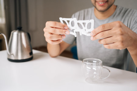 Cropped shot of smiling barista placing drip coffee bag onto glass cup, showcasing process of brewing freshly ground coffee with kettle in background. Concept of enjoying morning ritual.の写真素材