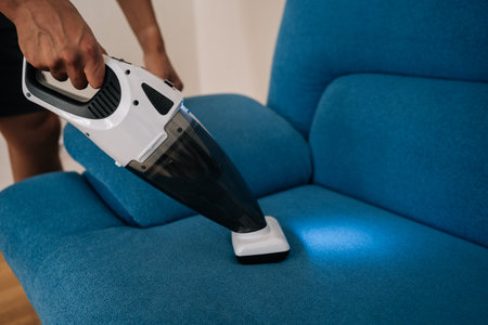 Close-up of cleaning service employee male using handheld vacuum cleaner to remove dust and dirt from blue sofa, emphasizing importance of cleanliness and hygiene in home maintenance.の写真素材