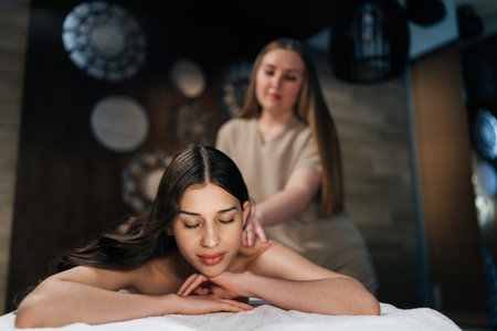 Closeup portrait of young woman lying with closed eyes in wellness center during massage therapy. Portrait of beautiful female client getting relaxing arm and shoulder massage in spa salon.の写真素材