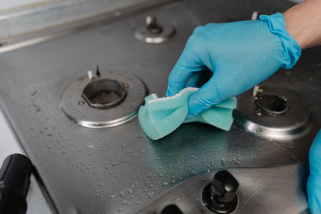 Professional housekeeper wearing blue gloves scrubbing dirty gas stove with sponge, applying cleaning solution for sparkling kitchen surfaces, close-up. Concept of cleanliness and housekeeping.の写真素材