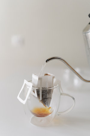 Pouring hot water from kettle into drip coffee bag nestled in glass cup, brewing fresh cup of coffee on white background, close-up. Process of making freshly brewed coffee at home during breakfast.の写真素材