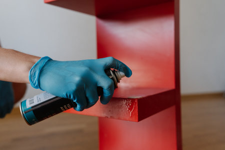 Male hand wearing blue glove applying scotch remover spray to dried adhesive stain on red shelf, performing thorough housekeeping at home, close-up. Concept of easy housekeeping domestic life.の写真素材