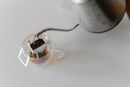 Close-up top view of pouring hot water from kettle into drip coffee bag nestled in glass cup, brewing fresh cup of coffee on white table. Process of making brewed coffee at home during breakfast.の写真素材