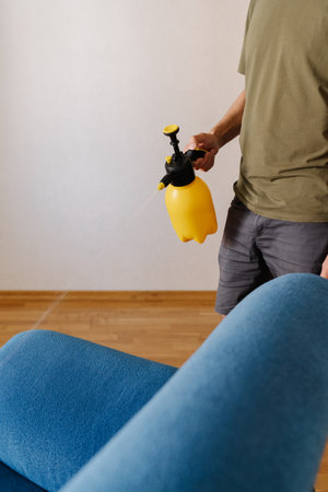 Househusband spraying water onto blue sofa using yellow sprayer, diligently cleaning furniture in cozy living room at home, ensuring hygiene and freshness, close-up. Concept of hygiene and cleanlinessの写真素材