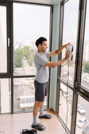 Full length vertical portrait of young man applying modern robotic device to large window in...