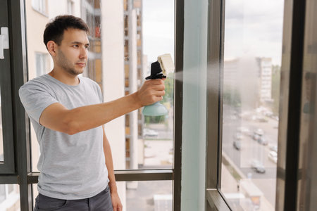 Janitor male spraying cleaning solution onto large window in high-rise building, preparing to wipe it clean with squeegee, ensuring spotless and sparkling finish for building occupants.の写真素材