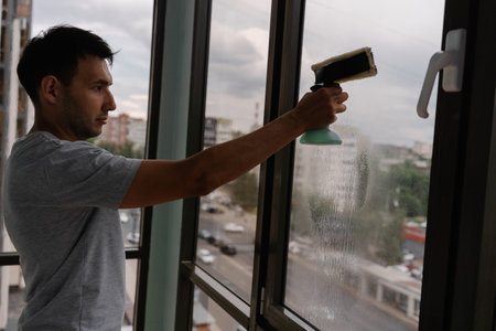 Maintenance worker male spraying cleaning solution on high-rise window, using squeegee for pristine, streak-free glass surface during professional building cleaning routine. Concept of cleanlinessの写真素材