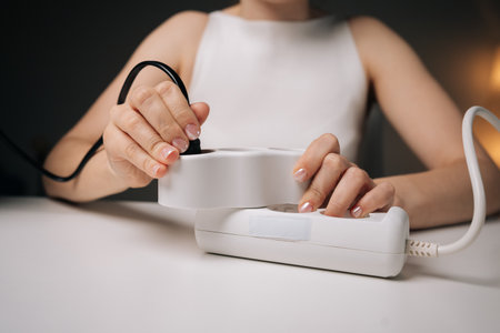 Close-up cropped shot of woman plugging black cable into white power strip on white table, with blurred background and soft lighting, creating sense of carefulness and precision.の写真素材