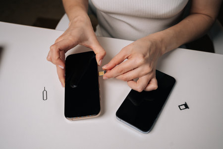 Close-up of young woman inserting sim card into new smartphone, with another phone and sim ejector tool nearby on white table, during mobile device setup process. Concept of modern technologyの写真素材