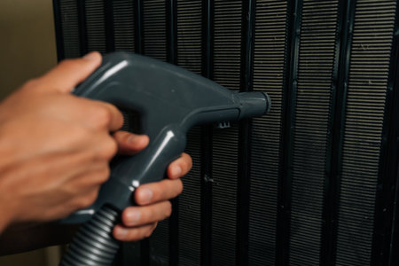 Man using vacuum cleaner attachment to remove dust and debris from condenser coils of refrigerator, ensuring efficient cooling and preventing malfunctions at home kitchen, close-up.の写真素材