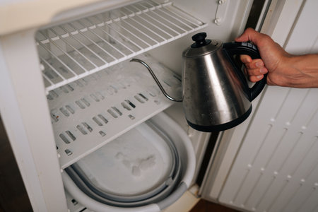 Close-up of unrecognizable person pouring hot water from kettle into refrigerator to melt ice and...