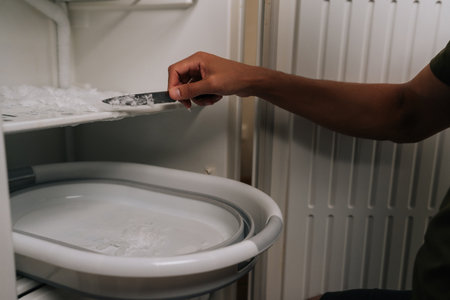 Close-up hands of unrecognizable man defrosting refrigerator, using knife to scrape off ice buildup...