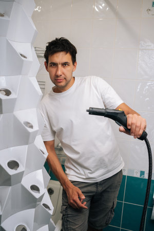 Vertical portrait of handsome man sanitizing vertical hydroponic tower garden with steam cleaner in bathroom, highlighting innovative indoor gardening and cleaning technique, posing looking at camera.の写真素材