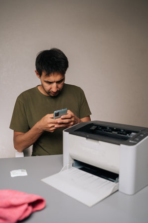 Focused man using smartphone to read instructions while repairing laser printer, with cleaning...