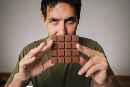 Happy young man holding chocolate bar in front of face, savoring delicious aroma, preparing to indulge in delightful sweet treat that promises pure enjoyment, sitting at table on isolated backgroundの写真素材