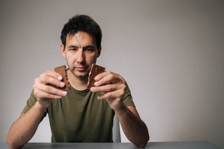 Portrait of young man sitting at a table, breaking chocolate bar into two pieces while contemplating which piece to indulge in first, reflecting on sweet temptation and craving for treat.の写真素材