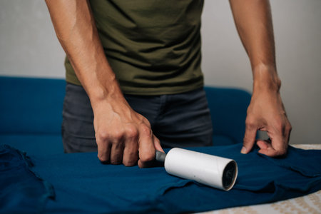 Close-up cropped shot of unrecognizable man using sticky lint roller to remove dust and lint from blue t-shirt on ironing board, preparing to iron and care for clothes at home.の写真素材
