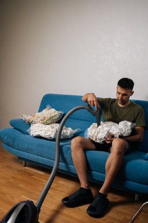 Portrait of man sitting on sofa using vacuum cleaner to suck air out of storage bag containing bedding, reducing storage space and protecting from dust and moisture. Concept of household chores.の写真素材