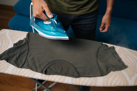 Close-up cropped shot of man carefully ironing dark green t-shirt on ironing board, using steam iron to remove wrinkles and achieve crisp at home. Concept of household chores and housework.