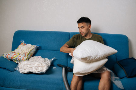 Young man sitting on sofa, using vacuum storage bag to organize and compress bedding, efficiently creating space-saving storage solution for tidy and clean home. Concept of space saving storage.の写真素材