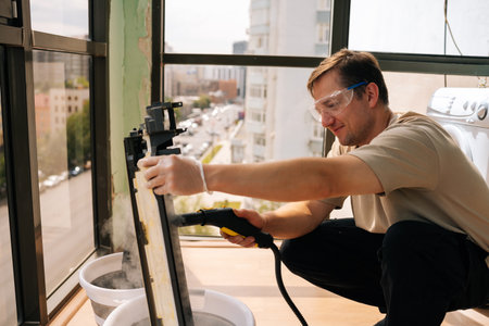 Professional cleaning service worker wearing safety glasses using steam cleaner to clean air conditioner filter in bucket of water, performing maintenance on HVAC system.の写真素材