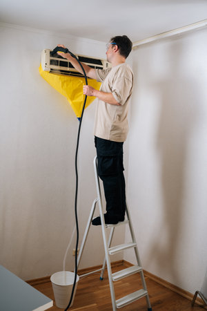 Remote view of professional technician in safety glasses standing on ladder, cleaning air conditioning unit with steam cleaner, yellow protective sheet covering wall surface to prevent water damageの写真素材
