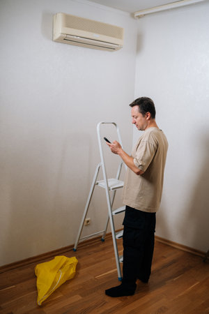 Vertical shot of technician male standing by ladder, using smartphone app to control and prepare for air conditioner maintenance, ensuring optimal cooling performance and indoor air quality.の写真素材
