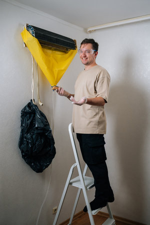 Vertical portrait of positive technician cleaning air conditioning unit, wearing protective gear, using sponge, yellow cover, smiling looking at camera. Concept of air condition hygiene.の写真素材