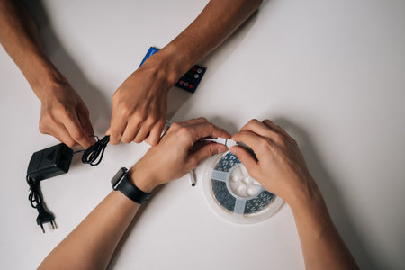Closeup cropped shot of two technicians connecting RGB led strip lights, configuring wireless controller for personalized ambient lighting in dark interior setting. Concept of modern technology.の写真素材