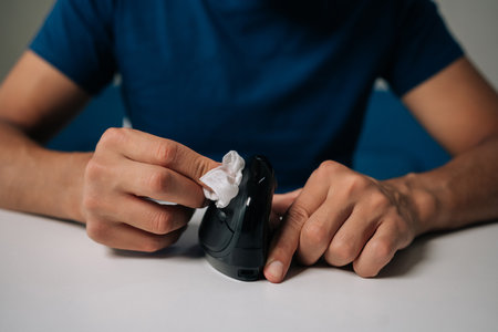 Cropped shot of unrecognizable man cleaning modern black vertical mouse with disinfectant wipe, promoting hygiene and preventing spread of germs, sitting at white table in workplace or home office.の写真素材