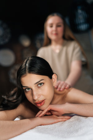 Vertical portrait of attarctive woman lying looking at camera in wellness center during massage therapy. Portrait of beautiful female client getting relaxing arm and shoulder massage in spa salon.の写真素材