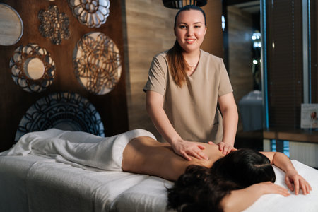 Portrait of smiling massage therapist performing deep massaging technique on female clients back in serene cozy spa environment, promoting relaxation and healing. Concept of rest with spa treatment.の写真素材