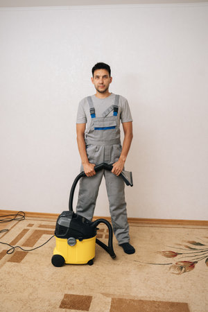 Full length vertical portrait of professional cleaning service worker in uniform holding washing vacuum cleaner, getting ready to clean carpet in living room posing on white background, looking cameraの写真素材