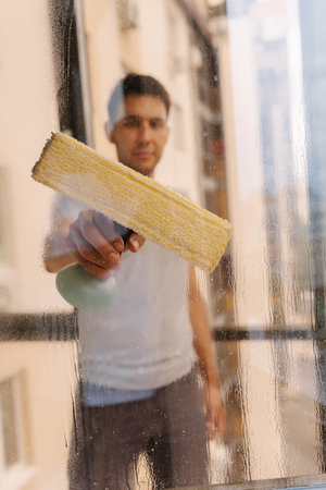 Vertical view from transparent window to young man cleaning large glass window using yellow squeegee, removing dirt with precise strokes and creating spotless surface in home environment.の写真素材