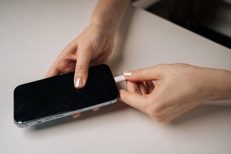 Close-up of unrecognizable woman connecting charging cable to modern mobile phone sitting at white table, capturing moment of charging smartphone and emphasizing interaction with technology, closeup.の写真素材