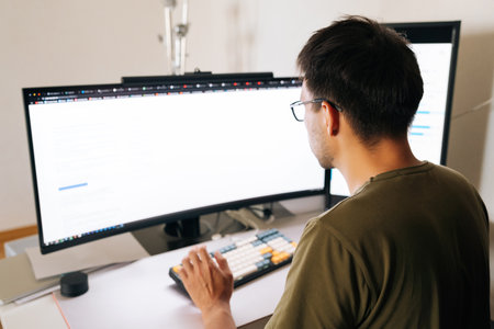View from shoulder of programmer in glasses concentrating on complex task, typing code on mechanical keyboard in home office, demonstrating modern work-from-home lifestyle. Concept of remote workplaceの写真素材