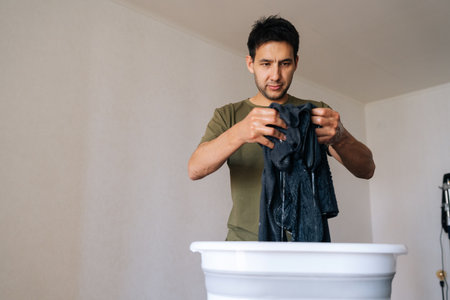 Portrait of focused young man squeezing water from black garment while hand washing clothes in white plastic basin at home, engaging in household chores and promoting sustainable living practices.の写真素材
