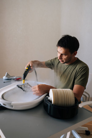 Vertical shot of focused man using steam cleaner to clean air humidifier filter in basin at home, ensuring regular maintenance for maintaining healthy indoor air quality. Concept of domestic work.の写真素材