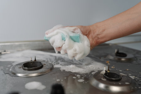 Homemaker hand squeezing soapy water from sponge during cleaning dirty gas stove, to removing grime,...
