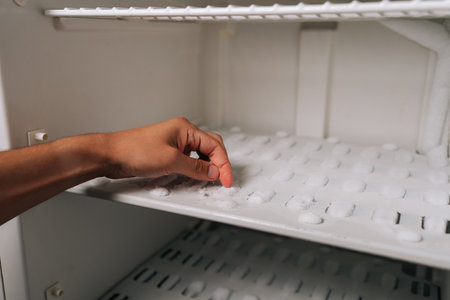 Fingertips pressing against thick frost layer inside refrigerator freezer, revealing heavy ice accumulation requiring urgent defrosting. Concept of need for defrosting due to excessive buildup.の写真素材